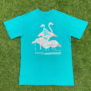 May include: A turquoise t-shirt featuring a flamingo design. The graphic includes pink flamingos with blue accents, and text that reads "NORTH CAROLINA ZOOLOGICAL SOCIETY COLLECTORS SERIES". The shirt is made of a soft material.