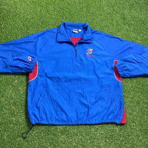 May include: A blue and red Kansas Jayhawks half-zip pullover jacket. The jacket has a stand-up collar, a zip closure, and the Kansas Jayhawks logo embroidered on the chest. The sleeves have red accents, and the bottom has a drawstring.