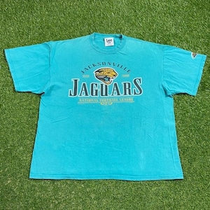 May include: Turquoise t-shirt featuring the Jacksonville Jaguars logo and text. The logo includes a jaguar head with the text "JAGUARS" below it. The shirt also has the text "NATIONAL FOOTBALL LEAGUE" and the NFL logo.