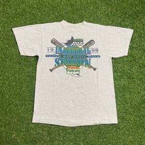 May include: A light gray t-shirt featuring a graphic for the Tampa Bay Devil Rays Inaugural Season in 1998. The design includes crossed baseball bats, the team name, and the Tropicana Field logo.