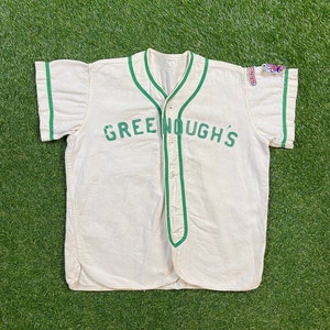 May include: White baseball jersey with green trim and the text "Greenough's" in green lettering.