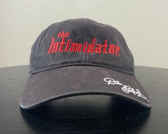 Könnte beinhalten: Eine schwarze Baseballkappe mit dem Schriftzug "The Intimidator" in roter Stickerei auf der Vorderseite. Die Kappe hat außerdem eine weiße Signatur an der Seite.