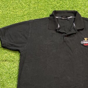 May include: Black polo shirt with a collared neckline and short sleeves. The Guinness logo is embroidered on the chest in white and red. The shirt is made of a soft, dark fabric.