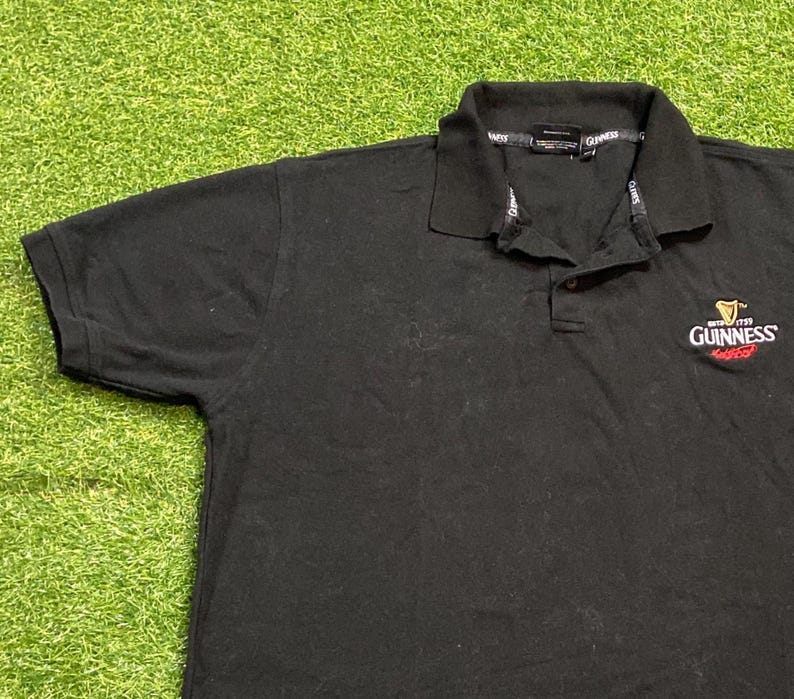 May include: Black polo shirt with a collared neckline and short sleeves. The Guinness logo is embroidered on the chest in white and red. The shirt is made of a soft, dark fabric.