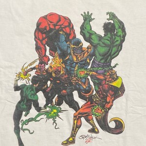 Op de afbeelding: Een wit T-shirt met een kleurrijke grafische print van verschillende superhelden, waaronder Hulk, Thanos en Green Lantern. De grafiek is gesigneerd door kunstenaar Gary Barker.
