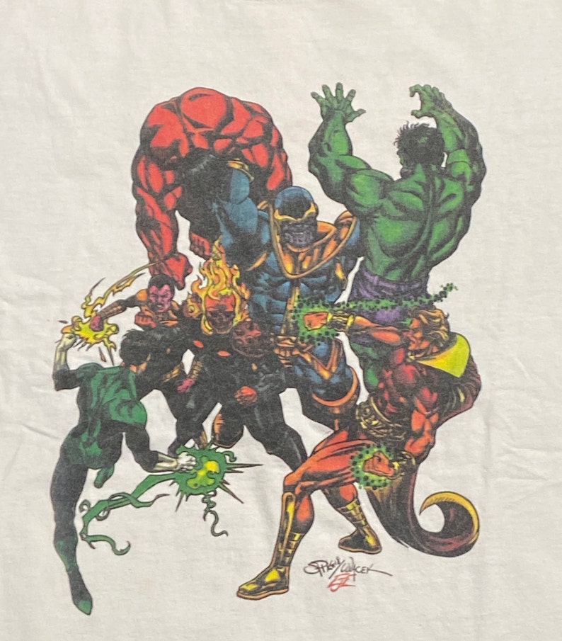 Op de afbeelding: Een wit T-shirt met een kleurrijke grafische print van verschillende superhelden, waaronder Hulk, Thanos en Green Lantern. De grafiek is gesigneerd door kunstenaar Gary Barker.