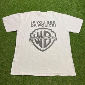 Könnte beinhalten: Weißes T-Shirt mit einem grauen Aufdruck. Der Aufdruck zeigt den Text "IF YOU SEE DA POLICE!" über einem schildförmigen Design mit "WARN A BROTHER" und "WB" darin. Das T-Shirt liegt flach auf einer grünen Oberfläche.