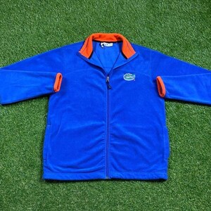 May include: Blue fleece jacket with orange trim and a small alligator logo on the chest. The jacket has a zipper closure and two pockets.
