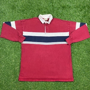 以下が含まれることがあります： 赤いベースに白、青、グレーのストライプが入った長袖のラグビーシャツ。シャツはボタンダウンカラーとボタン留めになっています。