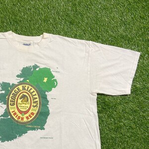 May include: White t-shirt with a green and red graphic of a map of Ireland and a George Killians Irish Red beer label. The label features a horse and the text "George Killians Irish Red. Premium Lager. Cheers. Brewed in the U.S.A. Enniscorthy."