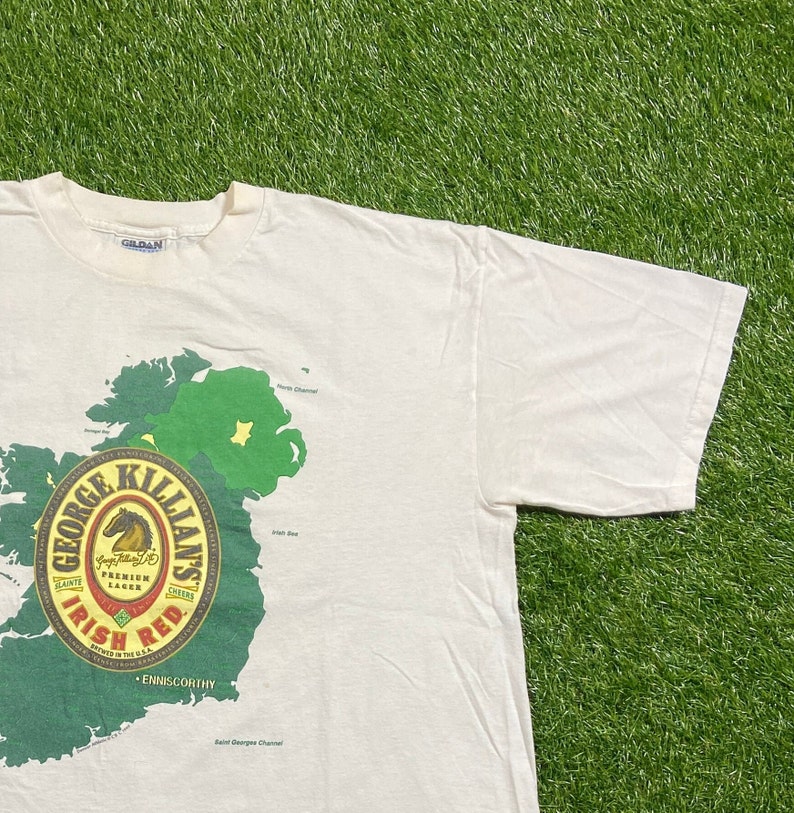 May include: White t-shirt with a green and red graphic of a map of Ireland and a George Killians Irish Red beer label. The label features a horse and the text "George Killians Irish Red. Premium Lager. Cheers. Brewed in the U.S.A. Enniscorthy."
