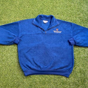 May include: A blue fleece pullover with a quarter-zip neckline. The pullover has a small orange and white logo with the text "1998 National Champions" on the chest. The cuffs and hem are a darker shade of blue.