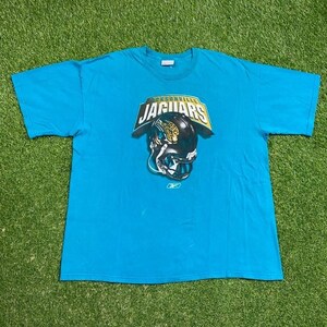 May include: A teal blue t-shirt with a Jacksonville Jaguars logo featuring a football helmet with a jaguar on it. The logo is in gold and black.