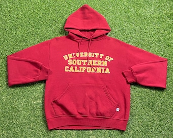 May include: A red hooded sweatshirt with the text "UNIVERSITY OF SOUTHERN CALIFORNIA" in yellow block letters. The sweatshirt has a front pocket and a drawstring hood. The Russell Athletic logo is visible on the lower right.