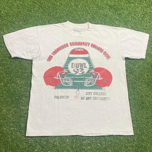 May include: White t-shirt with a football graphic. The design includes a football, two red helmets, and the text "San Francisco Community College Bowl" and "Bowl 93". Below, it reads "Palomar vs City College of San Francisco."