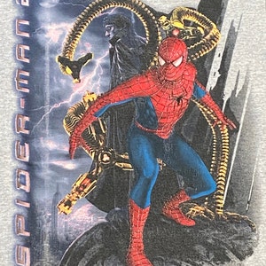 May include: A grey t-shirt featuring a graphic print of Spider-Man from the film Spider-Man 2. The image depicts Spider-Man in a red and blue suit with a black spider symbol on his chest. He is standing on a building with a cityscape in the background. The villain Doc Ock is in the background with his mechanical arms.