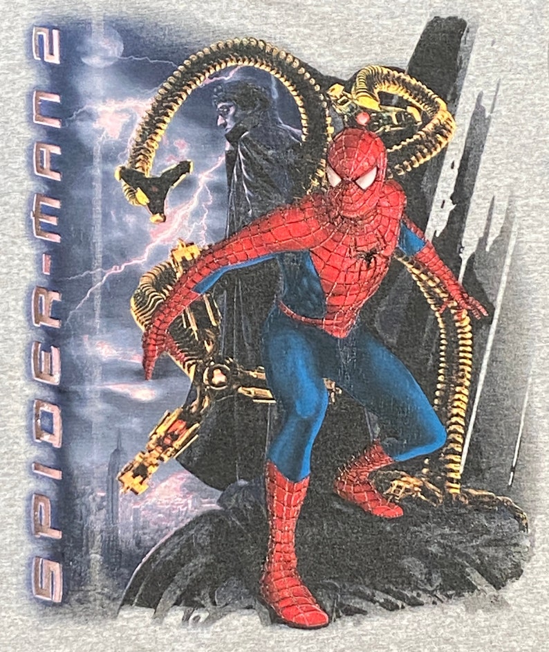 May include: A grey t-shirt featuring a graphic print of Spider-Man from the film Spider-Man 2. The image depicts Spider-Man in a red and blue suit with a black spider symbol on his chest. He is standing on a building with a cityscape in the background. The villain Doc Ock is in the background with his mechanical arms.