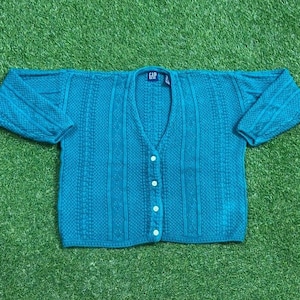 Könnte beinhalten: Türkisfarbene Strickjacke mit V-Ausschnitt und Knopfverschluss. Der Pullover hat ein strukturiertes Strickmuster und lange Ärmel. Das Markenetikett "GAP" ist am Ausschnitt sichtbar. Das Kleidungsstück liegt auf einer grünen Oberfläche.