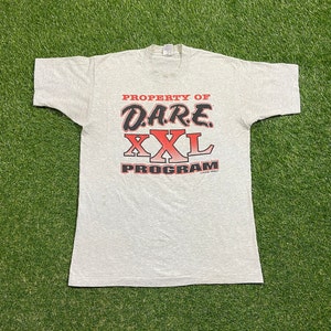 May include: A light gray t-shirt with the text "PROPERTY OF D.A.R.E. XXL PROGRAM" in red and black. The shirt is laid flat on a green grassy surface.