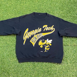 May include: Black sweatshirt featuring the words "Georgia Tech" in yellow script and a yellow and black cartoon bee. The words "YELLOW JACKETS" are also present. The sweatshirt is on a green surface.