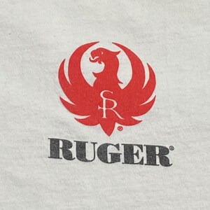 K&ouml;nnte beinhalten: Ein wei&szlig;es T-Shirt mit einem roten Ruger-Logo, das einen stilisierten Vogel mit den Buchstaben "SR" im Inneren zeigt. Das Wort "RUGER" ist in Schwarz unter dem Logo gedruckt.
