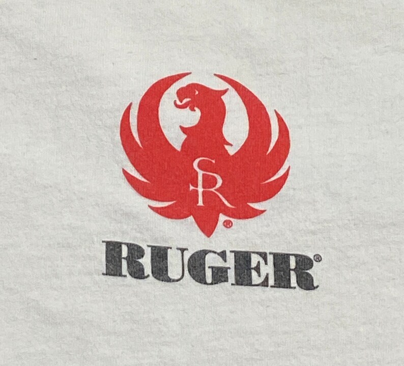 K&ouml;nnte beinhalten: Ein wei&szlig;es T-Shirt mit einem roten Ruger-Logo, das einen stilisierten Vogel mit den Buchstaben "SR" im Inneren zeigt. Das Wort "RUGER" ist in Schwarz unter dem Logo gedruckt.