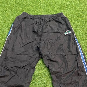 May include: Black nylon track pants featuring an elastic waistband, a small embroidered logo patch on the upper right side, and vertical blue and white piping accents running down the outer seams of both legs, creating a sporty appearance.