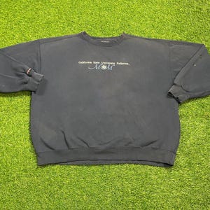Könnte beinhalten: Ein marineblaues Sweatshirt mit dem Text "California State University Fullerton" und dem Wort "MOM" in Schreibschrift. Das Sweatshirt liegt auf einer grünen Oberfläche.