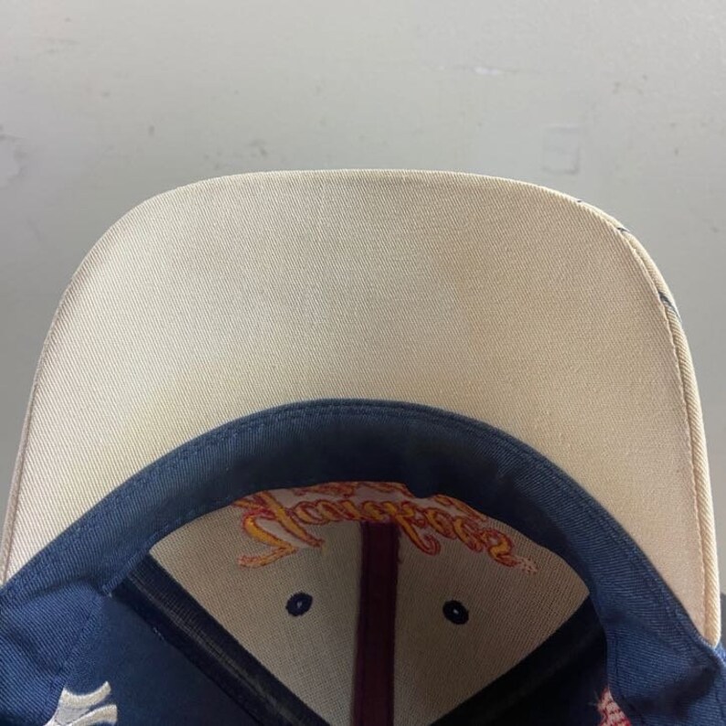 May include: A baseball cap with a tan brim and a navy blue crown. The underside of the brim features a colorful embroidered design. The cap has a curved brim and a structured fit.