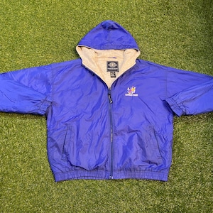 May include: A blue hooded jacket with a full zipper closure. The jacket has a beige lining in the hood and a small embroidered design with the words "Kansas Band". The jacket is made of a lightweight material.