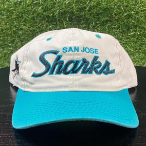 Può includere: Un cappellino da baseball bianco con visiera turchese e le parole "SAN JOSE Sharks" ricamate in turchese e nero. Un logo di squalo è sul lato. Il cappello è uno stile classico da baseball.