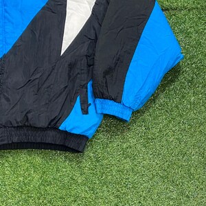 Peut inclure: Veste coupe-vent vintage noire, blanche et bleue. La veste pr&eacute;sente un design color-block avec une manche et des accents bleus. La veste est faite d'un mat&eacute;riau l&eacute;ger, possiblement en nylon.