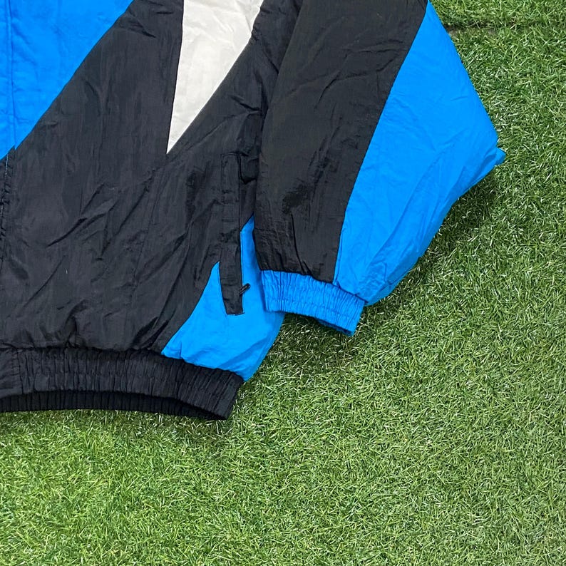 Peut inclure: Veste coupe-vent vintage noire, blanche et bleue. La veste pr&eacute;sente un design color-block avec une manche et des accents bleus. La veste est faite d'un mat&eacute;riau l&eacute;ger, possiblement en nylon.