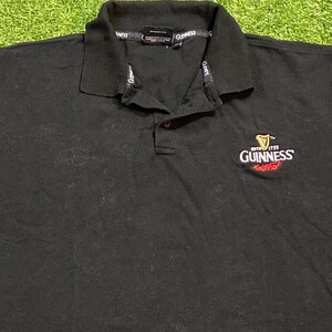 May include: Black polo shirt with a collared neckline and buttoned placket. The Guinness logo is embroidered on the left chest. The collar features the Guinness brand name.