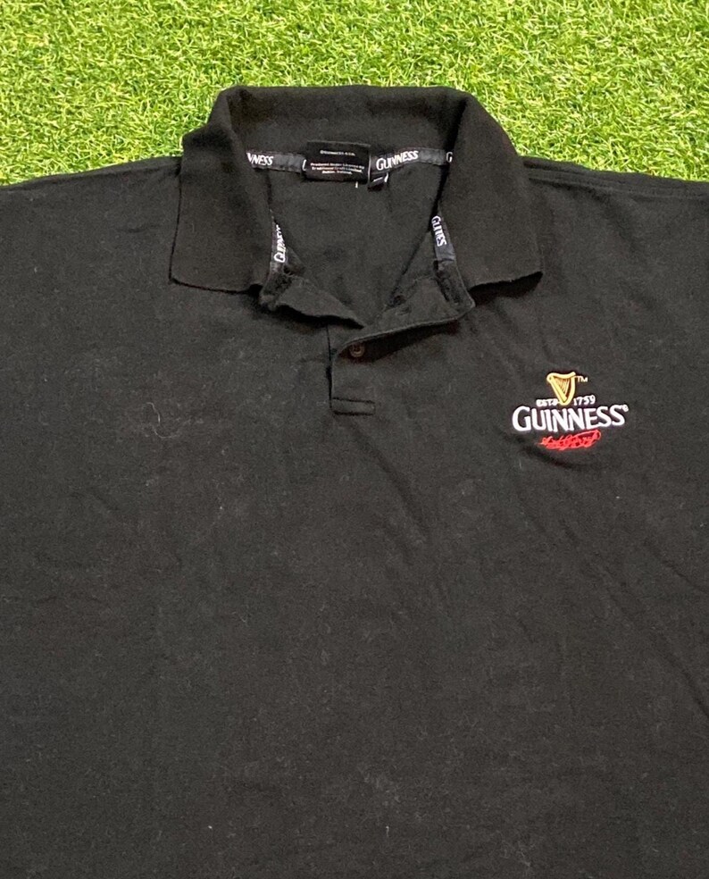 May include: Black polo shirt with a collared neckline and buttoned placket. The Guinness logo is embroidered on the left chest. The collar features the Guinness brand name.