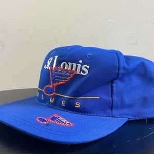K&ouml;nnte beinhalten: Eine blaue Baseballkappe mit einem wei&szlig;en gestickten St. Louis Blues Logo, das eine Musiknote und einen Fl&uuml;gel eines Hockeyspielers zeigt. Das Logo ist in Gold umrandet.