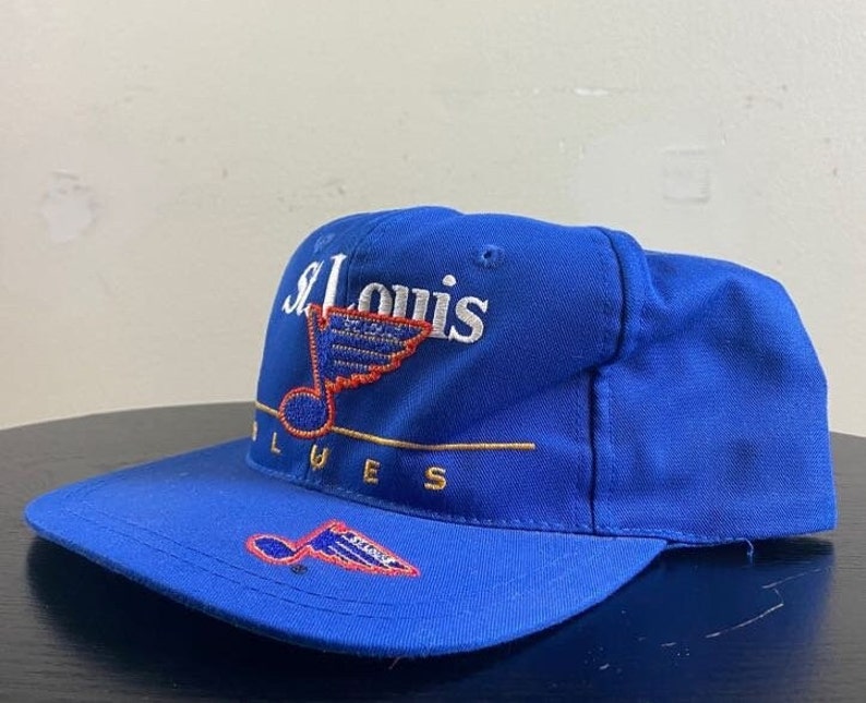 K&ouml;nnte beinhalten: Eine blaue Baseballkappe mit einem wei&szlig;en gestickten St. Louis Blues Logo, das eine Musiknote und einen Fl&uuml;gel eines Hockeyspielers zeigt. Das Logo ist in Gold umrandet.