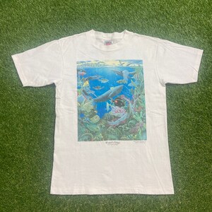 Könnte beinhalten: Weißes T-Shirt mit einem bunten Grafikdruck eines Wals und anderer Meeresbewohner in einem Korallenriff. Die Grafik ist mit "Water's Edge" betitelt.
