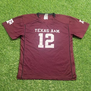 Peut inclure: Maillot de football bordeaux avec le texte "TEXAS A&M" et le numéro "12" en blanc. Le maillot a un col en V et des manches courtes. La lettre "E" est visible sur les manches.