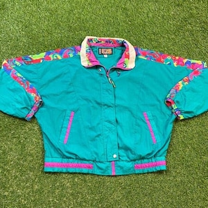 May include: Teal vintage windbreaker jacket with a colourful pattern on the shoulders and cuffs. The jacket has a white collar, pink accents, and a zip closure. The jacket has two front pockets with pink trim.