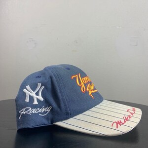 May include: A blue baseball cap with white pinstripes on the brim. The cap features the New York Yankees logo in white, the word "Racing" in white, and "Yankee Racing" in yellow and red script. The brim has "Mike Dunn" in red script.