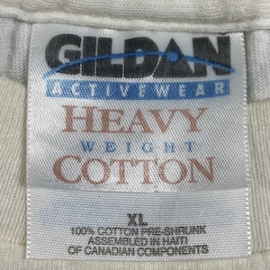 May include: White Gildan brand t-shirt tag with the text "Heavy Weight Cotton" and "100% Cotton Pre-Shrunk Assembled in Haiti of Canadian Components". The tag also includes the size "XL".