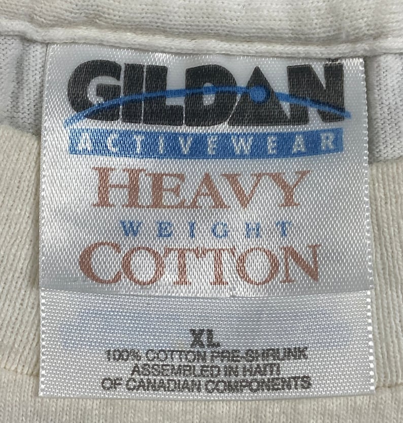May include: White Gildan brand t-shirt tag with the text "Heavy Weight Cotton" and "100% Cotton Pre-Shrunk Assembled in Haiti of Canadian Components". The tag also includes the size "XL".