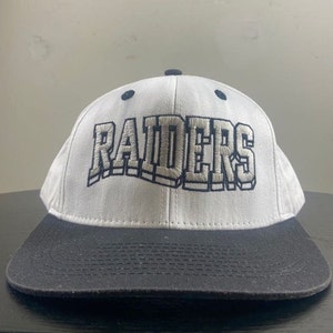 May include: White and black baseball cap with the word "RAIDERS" embroidered in a 3D style on the front.