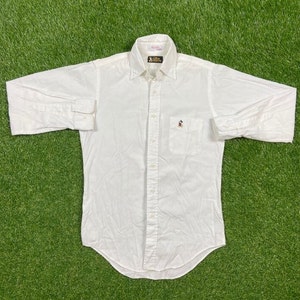 Könnte beinhalten: Weißes, langärmliges Hemd mit Knöpfen und einem kleinen Mickey-Mouse-Logo auf der linken Brusttasche. Das Hemd hat einen spitzen Kragen und einen Knopfmanschetten.