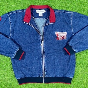 May include: A blue denim jacket with a red lining and a zipper closure. The jacket has a red embroidered logo on the left chest that says "Sooners" and features a wagon with a horse pulling it. The jacket has a black and red trim around the cuffs and bottom.