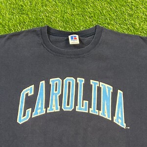 May include: A navy blue t-shirt with the word "CAROLINA" printed in light blue with a white outline. The shirt has a small tag on the front with the letter "R" in a circle.