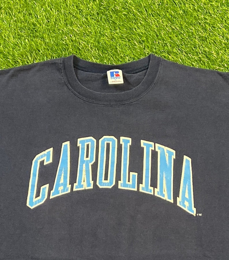 May include: A navy blue t-shirt with the word "CAROLINA" printed in light blue with a white outline. The shirt has a small tag on the front with the letter "R" in a circle.