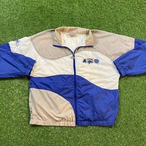 以下が含まれることがあります： 白、青、グレーのウィンドブレーカー。前面に「Orlando Magic」の文字が入っています。ジッパー開閉式で、ドローストリングフード付きです。