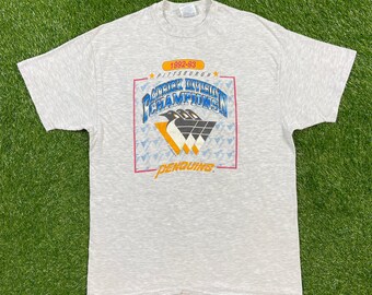 May include: A gray t-shirt with a graphic of the Pittsburgh Penguins hockey team. The graphic features the team's logo, the text "1992-93 Pittsburgh Patrick Division Champions", and the words "Penguins" in a banner below the logo.